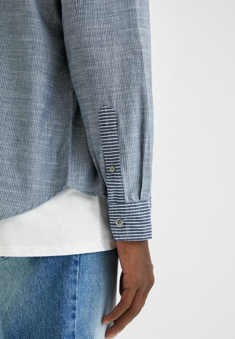 Blau-weiß gestreiftes Langarmhemd mit geknöpften Manschetten. Besitzt einen strukturierten Stoff und einen abgerundeten Saum, getragen über einem weißen T-Shirt.