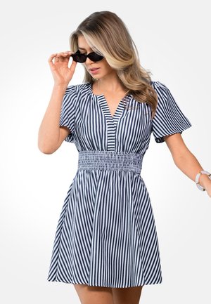 Kobieta poprawiająca czarne okulary przeciwsłoneczne, ubrana w sukienkę z krótkim rękawem w pionowe paski w kolorze granatowym i białym, z marszczoną talią i rozkloszowanym dołem.