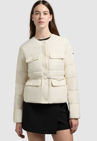 Donna che indossa una giacca trapuntata color crema con bottoni a pressione e tasche con pattina frontali, abbinata a una gonna asimmetrica nera.