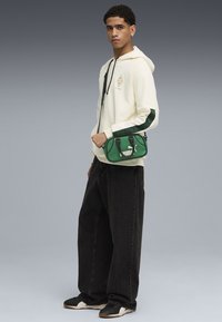 Tânăr îmbrăcat într-o hanorac crem cu dungi verzi, pantaloni largi negri, pantofi sport negri și purtând o geantă mică Puma verde crossbody, stând pe un fundal gri.