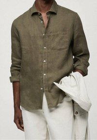 Olijfgroen linnen overhemd met knopen, voorzien van een borstzak, opgerolde mouwen en lichte broeken, vastgehouden met een licht jack dat over de arm is gedrapeerd.