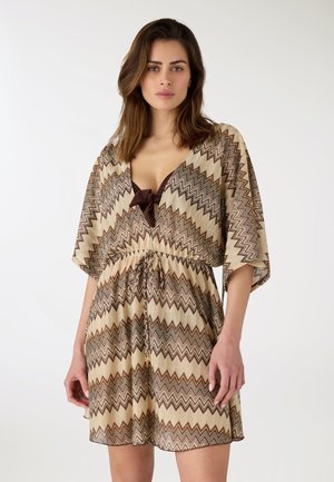 Donna che indossa un vestito marrone con motivo a zigzag, maniche larghe e vita con coulisse, in piedi davanti a uno sfondo chiaro uniforme.