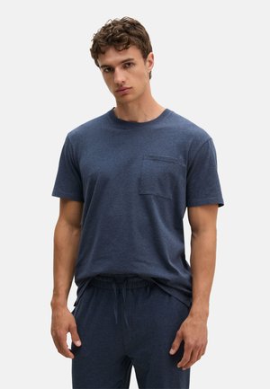 Jonge man draagt een donkerblauw T-shirt met korte mouwen en borstzakje en bijpassende broek met trekkoord, staand tegen een effen achtergrond.