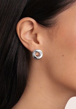 Orecchini circolari in metallo argento con un ritaglio centrale, superficie lucida e texture liscia, indossati all'orecchio di un modello.