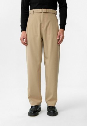 Khaki broek met een ontspannen pasvorm, een vlakke voorkant en een riem met een zilveren gesp. De stof lijkt glad met een lichte glans. Gecombineerd met zwarte schoenen.