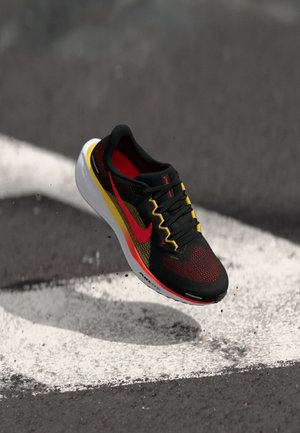 Schwarzer, roter und gelber Sportschuh mit weißer Sohle erscheint in der Luft über Asphalt mit weißen Markierungen.