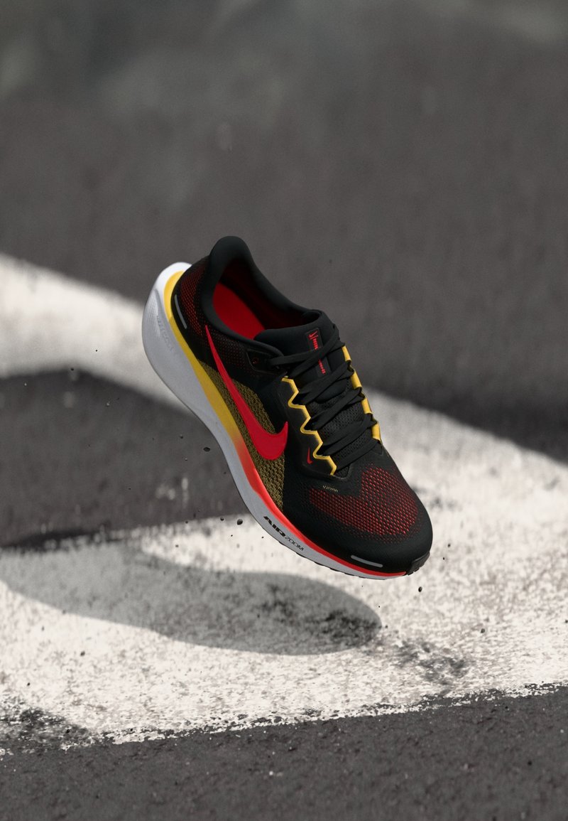 Chaussure de sport noire, rouge et jaune avec une semelle blanche apparaît en l'air au-dessus d'un bitume avec des marquages blancs.