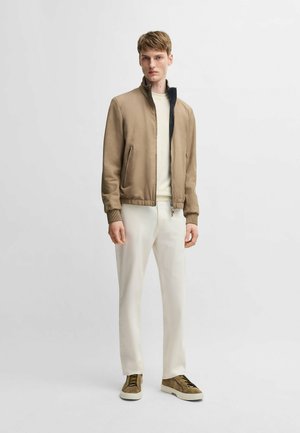 Brown bomber jacket with a high collar, white interior, ribbed cuffs, and two side pockets. Worn with white trousers and brown sneakers.