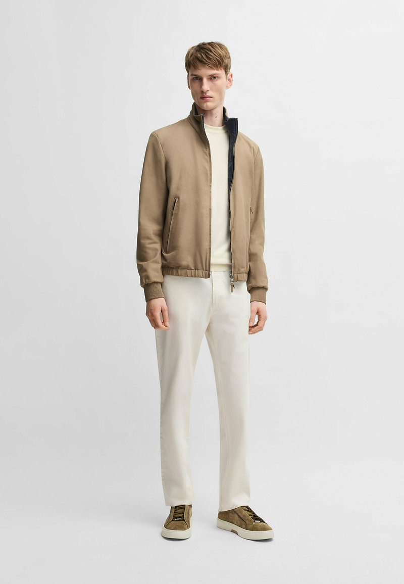 Giacca bomber marrone con colletto alto, interno bianco, polsini a coste e due tasche laterali. Indossata con pantaloni bianchi e sneakers marroni.