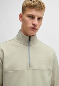 Felpa leggera color beige chiaro con zip a metà, realizzata in tessuto testurizzato e dotata di un logo sottile sul petto. Presenta un colletto alto e una zip grigia.