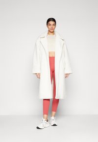 Cappotto bianco e morbido con un ampio colletto, indossato sopra un top corto beige e leggings rosa, abbinato a sneaker bianche. Design semplice e lineare.