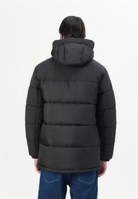 Giacca puffer nera con cappuccio staccabile, caratterizzata da un motivo trapuntato, polsini elastici e un retro leggermente più lungo per una maggiore copertura.