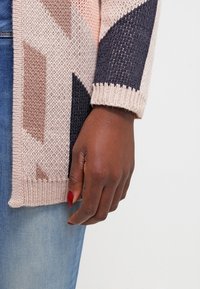 Stickad cardigan med geometriska mönster i beige, rosa och marinblått. Har en texturerad finish och ribbade manschetter. Matchas med blå denim.