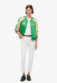 Veste bomber en satin vert et beige avec des détails brodés, poignets et ourlet côtelés, portée avec une chemise blanche et un pantalon blanc. Chaussures noires.