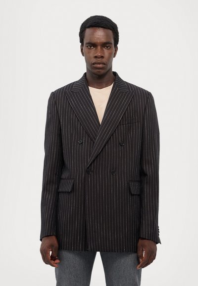 Dobbeltbrystet sort blazer med hvide striber, med notched lapels og sidelommer, lavet af tekstureret stof. Neutral skjorte under.