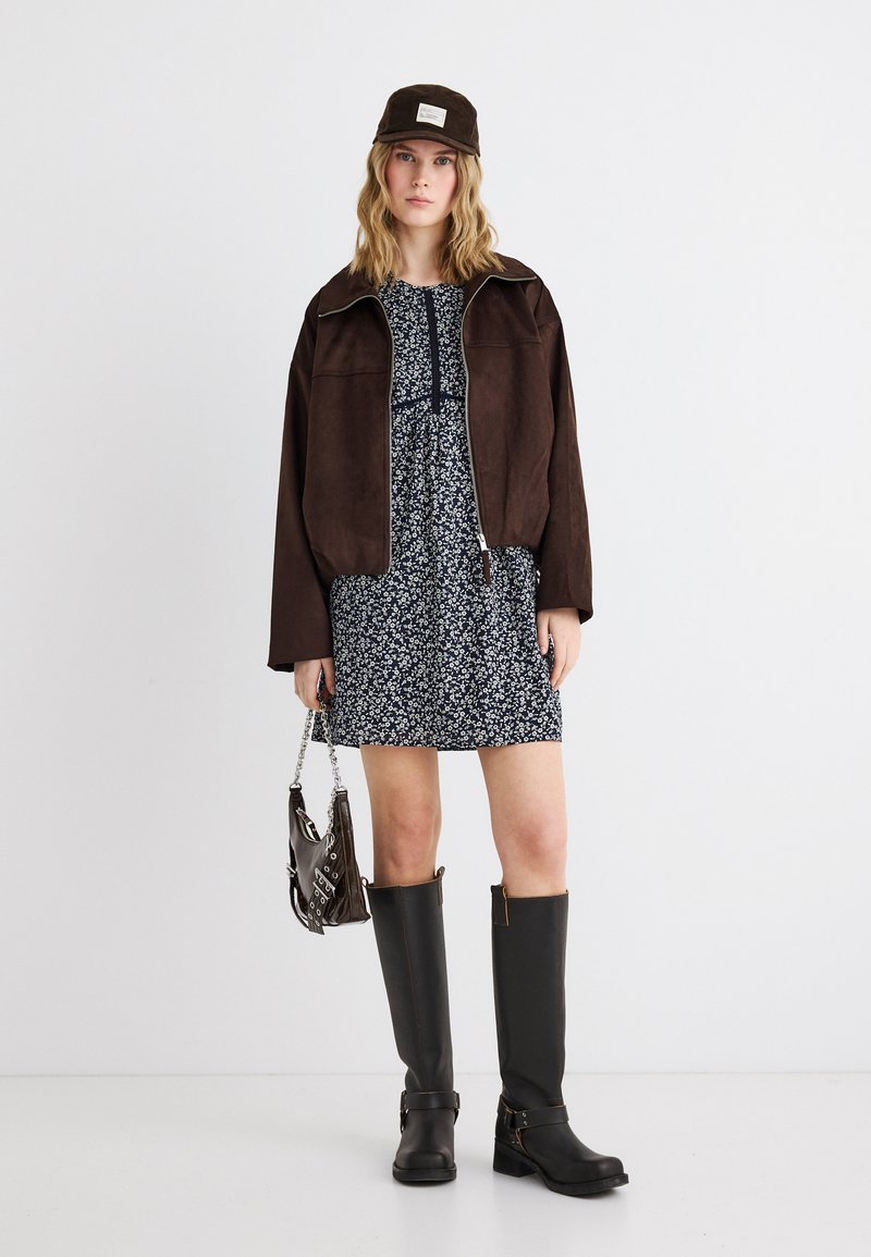 Γυναίκα που φοράει σκούρο καφέ μπουφάν και σκουφάκι, μαύρο φόρεμα με λουλούδια, μαύρες μπότες μέχρι το γόνατο και κρατά μικρό καφέ τσάντα.