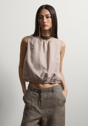 Jeune femme aux longs cheveux lisses portant un top beige sans manches et un pantalon marron taille haute, debout les mains dans les poches.