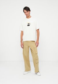 White short-sleeve t-shirt with a graphic of a coffee pot and text, paired with beige cargo pants featuring multiple pockets and a relaxed fit.