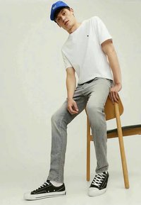 Jovem usando um boné azul, uma t-shirt branca, jeans cinzentos e sapatilhas pretas, sentado numa cadeira de madeira contra um fundo simples.
