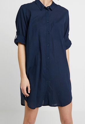 Granatowa sukienka koszulowa z podwiniętymi rękawami, kieszenią na piersi i subtelnymi pionowymi paskami, noszona na długość do połowy uda.