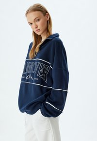 Sudadera de forro polar azul marino con cuello alto, cierre de media cremallera, franjas de acento blancas y gráfico "HANOVER" en el pecho.