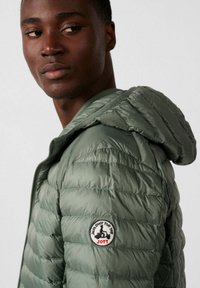 Giacca a vento imbottita verde oliva con motivo trapuntato, cappuccio e logo ricamato sulla manica sinistra. Realizzata in materiale leggero e resistente all'acqua.