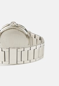 Montre en acier inoxydable argenté avec un boîtier arrondi, une couronne texturée et un bracelet à maillons ; le dos présente une marque gravée.