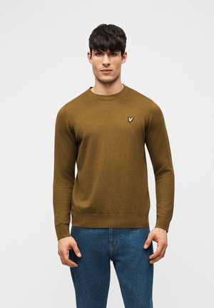 Homme portant un pull brun à manches longues avec un petit logo d'oiseau jaune sur la poitrine et un jean bleu, debout devant un fond uni.