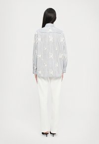 Chemise à rayures bleu et blanc avec des détails en dentelle florale ; manches longues, coupe décontractée. Associée à un pantalon blanc sur mesure et des talons noirs.