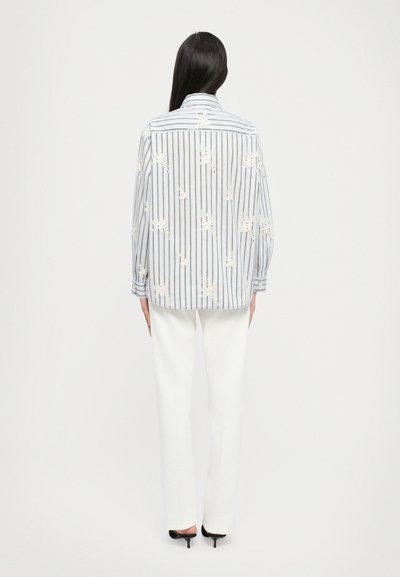 Striped button-up shirt in blue and white with floral lace details; long sleeves, relaxed fit. Paired with white tailored trousers and black heels.