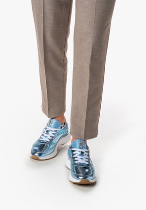 Baskets de sport en bleu métallique brillant avec des lacets blancs, surface texturée et une semelle blanche épaisse. Portées avec un pantalon beige sur mesure.