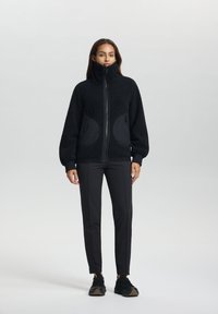 Zwarte fleecejas met hoge kraag, gestructureerde stof, ritssluiting en twee grote ronde zakken. Gepaard met zwarte slim-fit broek en sneakers.