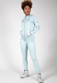 Ensemble de sport bleu clair comprenant un sweat à capuche zippé et un pantalon ajusté assorti. Tissu lisse et extensible ; dispose de poches latérales et d'une capuche.