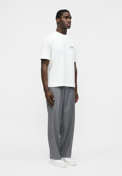 Homme portant un T-shirt blanc à manches courtes avec un petit logo et un pantalon gris ample, debout de profil sur un fond uni.