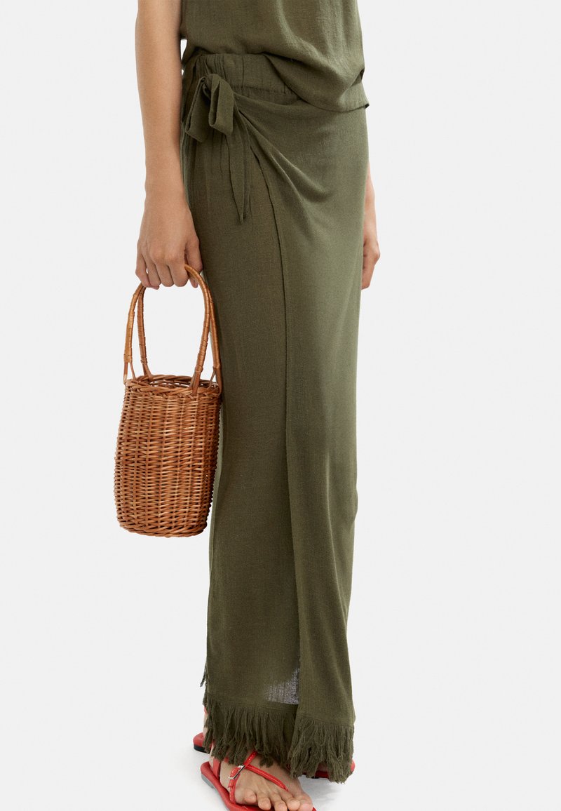 Oliwkowa spódnica z bocznym wiązaniem, tkaniną o fakturze wplecionej, z frędzlami na dole; połączona z okrągłą wiklinową torbą i czerwonymi sandałami.