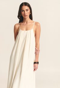 Robe maxi sans manches de couleur crème avec une coupe fluide, dotée de fines bretelles et de plis verticaux. Tissu lisse et léger.