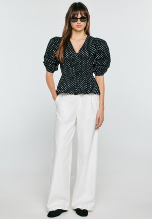 Kobieta ubrana w czarną bluzkę w groszki z bufkami na rękawach, białe szerokie spodnie, czarne baleriny i czarne okulary przeciwsłoneczne, stoi z rękami w kieszeniach.