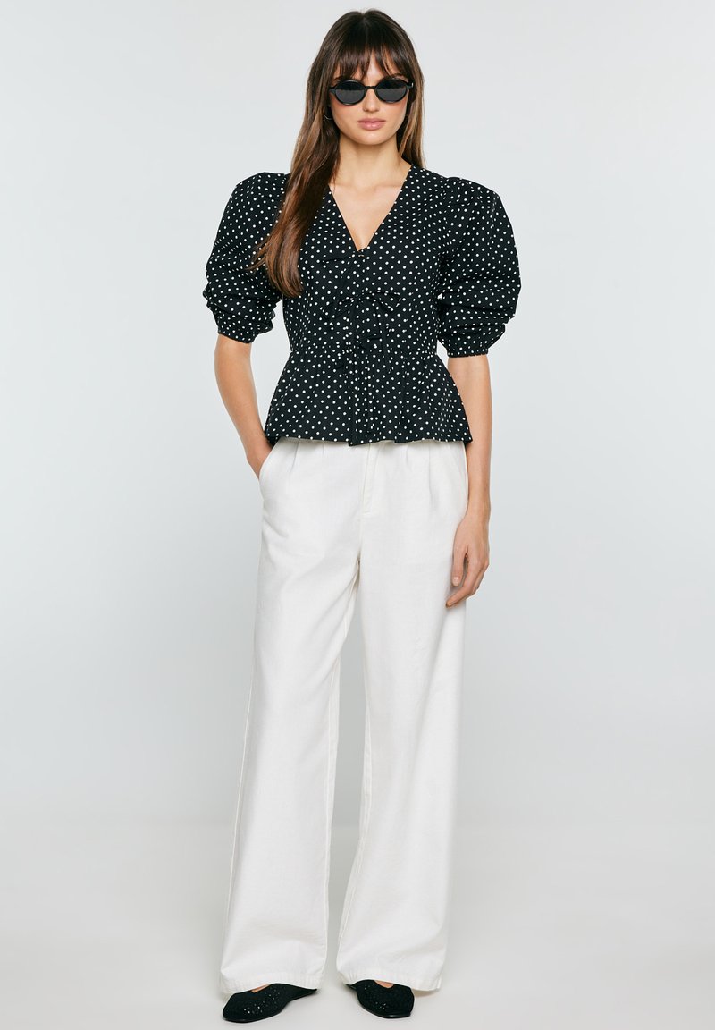 Mujer con blusa negra de lunares con mangas abullonadas, pantalones blancos de pierna ancha, zapatos planos negros y gafas de sol negras, de pie con las manos en los bolsillos.