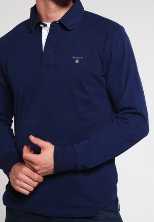 Mann trägt ein dunkelblaues Langarm-Poloshirt mit kleinem GANT-Logo auf der Brust, er richtet mit der rechten Hand seinen linken Ärmel.