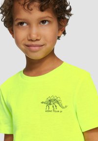 Neon gele t-shirt met een groene dinosaurus-skeletafbeelding en de tekst "#DINO TEAM UP" op de voorkant. Korte mouwen, ronde hals.