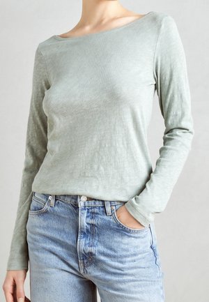 Personne portant une chemise à manches longues vert clair rentrée dans un jean taille haute bleu clair avec une main dans la poche.