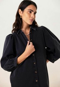 Dunkelblaue Jeanshemd mit Knopfleiste, gepufften Ärmeln und klassischem Kragen. Besitzt eine glatte Textur und dezente Nähte.