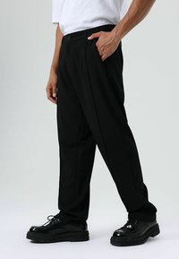 Zwarte, losse broek met knoopsluiting, voorzien van zijzakken en een gladde textuur, gecombineerd met glanzende zwarte schoenen.