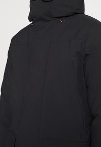 Zwarte geïsoleerde jas met een hoge kraag, verstelbare capuchon en zijzakken. Gladde textuur met contrasterende hardware accenten.