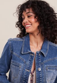 Veste en denim bleu moyen avec un col classique, fermeture à boutons et deux poches poitrine. Présente une finition légèrement délavée et une coupe ajustée.