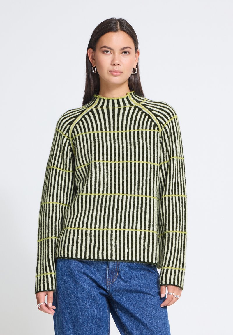 Femme aux cheveux raides foncés portant un pull rayé noir et blanc avec de fines lignes horizontales jaunes et un jean bleu, debout devant un fond blanc.
