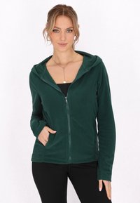 Felpa zip-up verde scuro in microfibra con una consistenza morbida, tasche anteriori e cappuccio; indossata sopra una maglietta nera. Design minimalista con linee semplici.