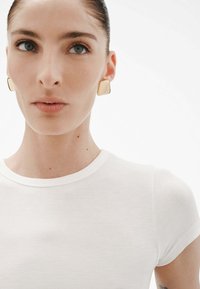 Camiseta blanca de canalé con mangas cortas, que presenta un cuello redondo. La modelo lleva puestos pendientes cuadrados de oro y tiene una piel suave.