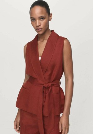 Una donna in un gilet senza maniche color ruggine con cintura e pantaloni abbinati, di fronte a uno sfondo chiaro, che guarda avanti con un'espressione seria.