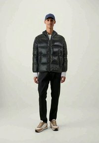 Giacca a vento in grigio scuro con cerniera, due tasche frontali e cappuccio, abbinata a pantaloni neri e sneakers beige. Sfondo chiaro.